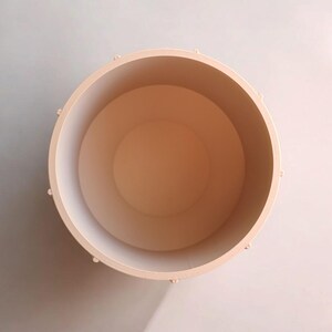 May include: A top-down view of a light peach-colored cylindrical container. The container has a smooth interior and exterior, with small, evenly spaced raised details around the rim. The inside is a slightly darker shade of peach.