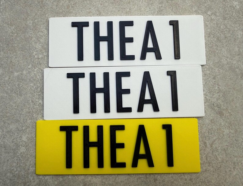 Pu&ograve; includere: Tre targhe rettangolari. Due sono bianche e una &egrave; gialla. Ogni targa riporta la scritta "THEA 1" in lettere nere e in grassetto. Le targhe sono disposte orizzontalmente, con quella gialla in basso.