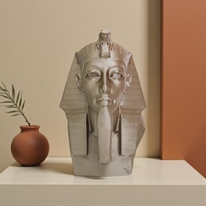 May include: A silver-colored bust of an Egyptian pharaoh, featuring a detailed headdress and serene facial expression. The sculpture is displayed on a white surface, with a small terracotta vase and plant in the background.