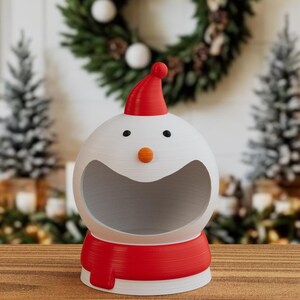 May include: A 3D-printed snowman-shaped container with a red hat and scarf. The snowman has a large open mouth, black eyes, and an orange nose. The container is white with red accents, ideal for festive decorations.