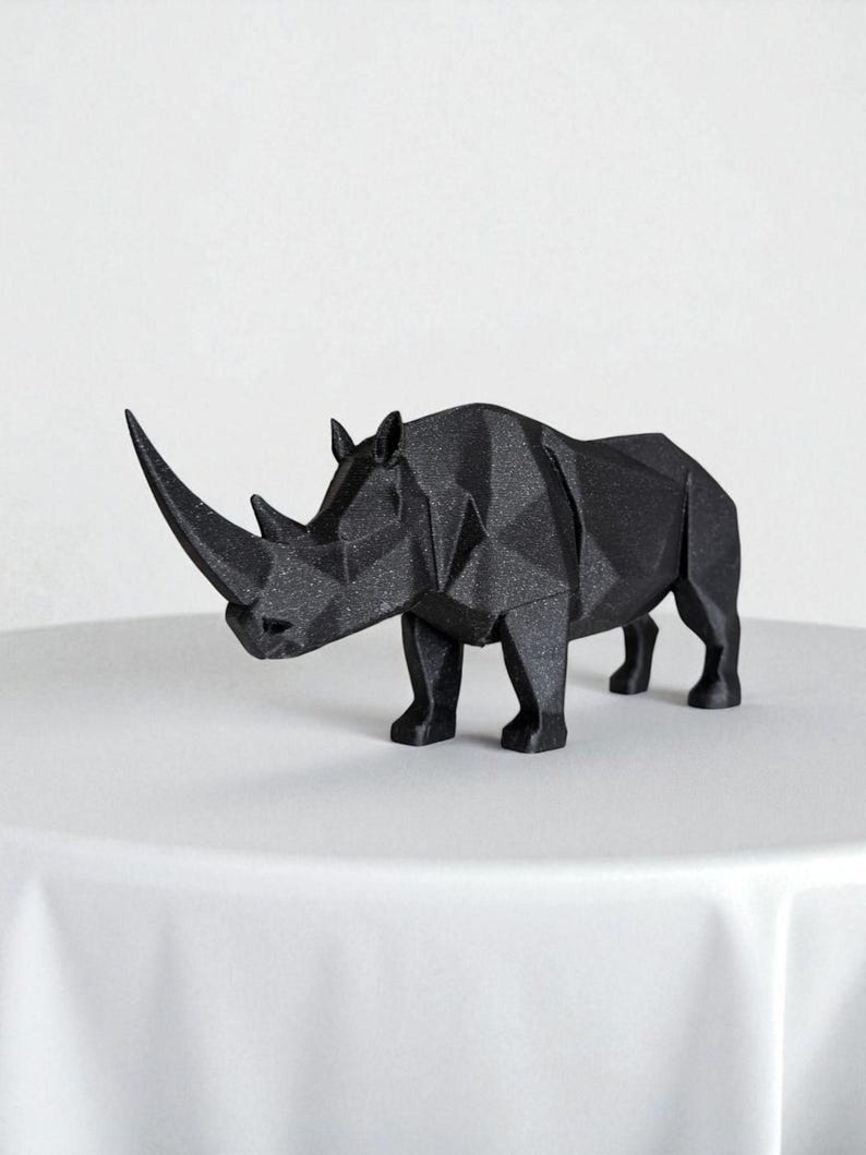 May include: A black, geometric rhino sculpture with a textured surface. The rhino is standing on a white surface. The sculpture has a large horn and is a modern decorative piece.