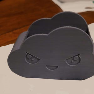 May include: A dark gray cloud-shaped desk organizer with an angry face. The organizer has a slot for holding items and is made of a textured material. The design features two eyes and a smiling mouth.