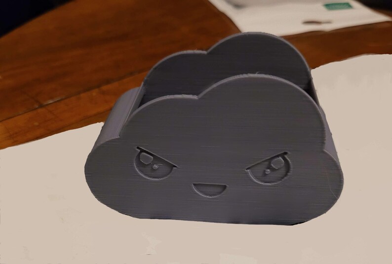May include: A dark gray cloud-shaped desk organizer with an angry face. The organizer has a slot for holding items and is made of a textured material. The design features two eyes and a smiling mouth.