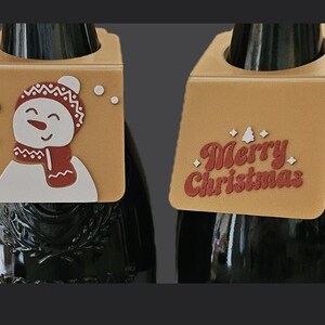 May include: Two Christmas-themed wine bottle tags. One tag features a snowman with a red scarf and hat, and the other says "Merry Christmas" in red lettering. Both tags are tan with a hole for the bottle neck.