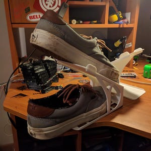 May include: Two gray canvas sneakers with brown leather accents are displayed on a white 3D-printed shoe rack. The shoes are worn and show signs of use. The rack is on a wooden desk.