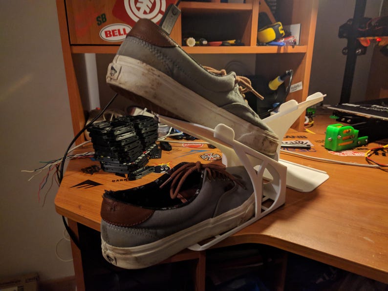 May include: Two gray canvas sneakers with brown leather accents are displayed on a white 3D-printed shoe rack. The shoes are worn and show signs of use. The rack is on a wooden desk.