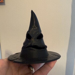 Op de afbeelding: Een zwarte, 3D-geprinte Sorteerhoed met een brede rand en een puntige top. De hoed heeft een gezichtachtig ontwerp met oog- en mondopeningen. De hoed wordt in een hand gehouden.