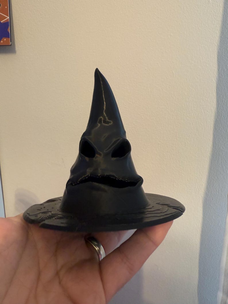 Op de afbeelding: Een zwarte, 3D-geprinte Sorteerhoed met een brede rand en een puntige top. De hoed heeft een gezichtachtig ontwerp met oog- en mondopeningen. De hoed wordt in een hand gehouden.