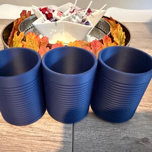 Pode incluir: Tr&ecirc;s canecas cil&iacute;ndricas azul-marinho com detalhes de ranhuras horizontais. As canecas est&atilde;o dispostas em fila sobre uma superf&iacute;cie de madeira. No fundo, uma bandeja decorativa cont&eacute;m folhas de outono e uma tigela de doces.
