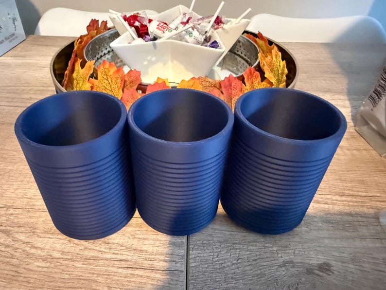 Pode incluir: Tr&ecirc;s canecas cil&iacute;ndricas azul-marinho com detalhes de ranhuras horizontais. As canecas est&atilde;o dispostas em fila sobre uma superf&iacute;cie de madeira. No fundo, uma bandeja decorativa cont&eacute;m folhas de outono e uma tigela de doces.