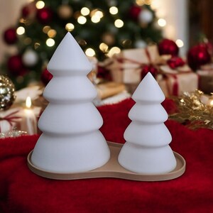 May include: Two white ceramic Christmas tree shaped candles on a brown tray. The candles are on a red surface.