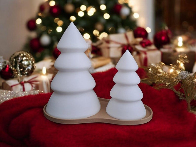 May include: Two white ceramic Christmas tree shaped candles on a brown tray. The candles are on a red surface.