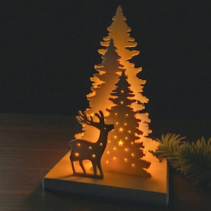 May include: A wooden Christmas decoration featuring a deer silhouette and a layered tree design. The tree has star-shaped cutouts that illuminate with a warm glow. The base is a rectangular block, creating a festive holiday scene.