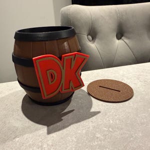 May include: A brown barrel-shaped container with black bands and a red and gold "DK" logo. A brown, round lid with a slot sits beside the container. The container is on a gray surface.