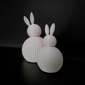 May include: Two white bunny figurines with rounded bodies and long ears. One has a ribbed texture on the lower body, while the other is smooth. The figurines are set against a black background, creating a minimalist aesthetic.