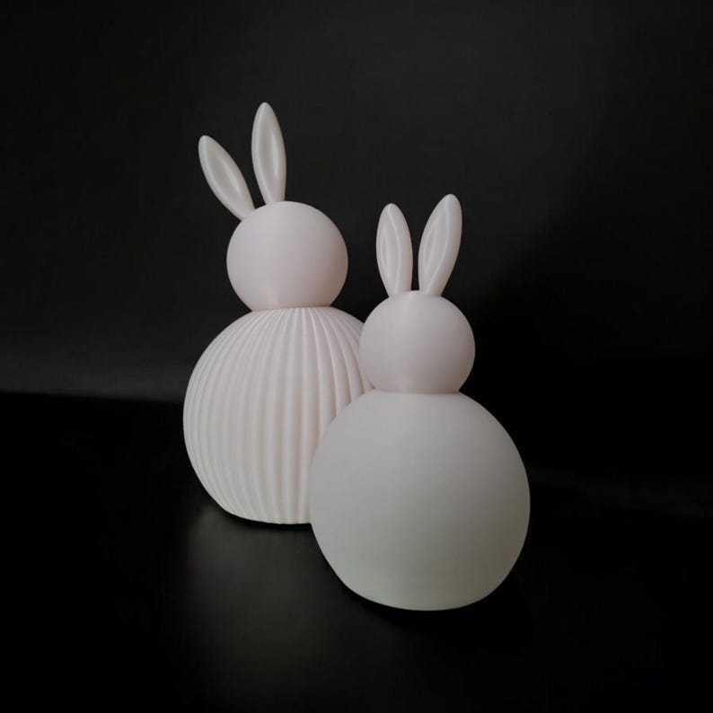 May include: Two white bunny figurines with rounded bodies and long ears. One has a ribbed texture on the lower body, while the other is smooth. The figurines are set against a black background, creating a minimalist aesthetic.