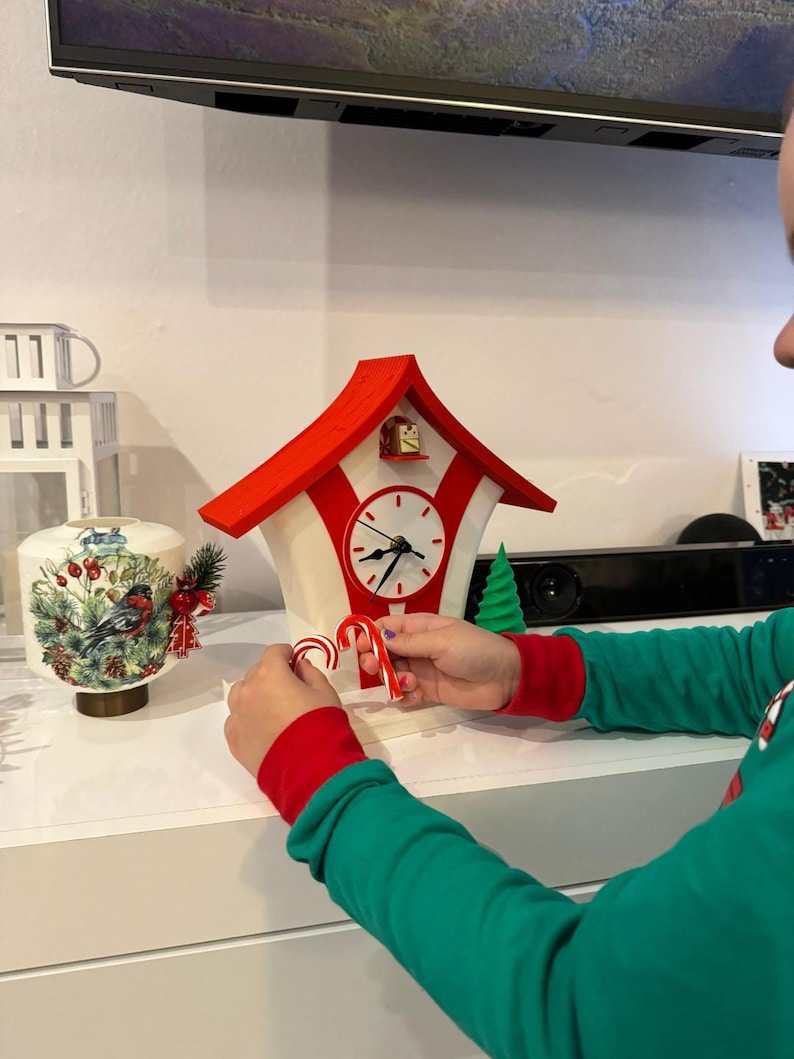 May include: A red and white decorative cuckoo clock with a birdhouse design. The clock has a round face with black hands and a pendulum. A person is holding two candy canes in front of the clock. A Christmas theme.