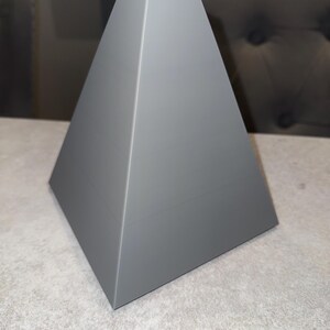 May include: A gray, pyramid-shaped decorative object with a square opening at the top. The sides slope inward, creating a geometric design. The object is made of a solid material and sits on a light-colored surface.