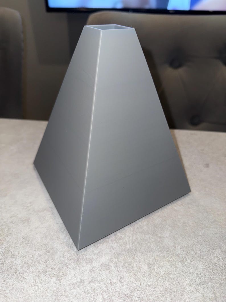 May include: A gray, pyramid-shaped decorative object with a square opening at the top. The sides slope inward, creating a geometric design. The object is made of a solid material and sits on a light-colored surface.