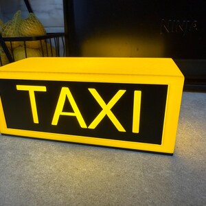 May include: A yellow rectangular taxi sign with the word "TAXI" in bold, illuminated letters. The sign has a black background and is lit from within, creating a bright, eye-catching display. The sign is on a gray surface.