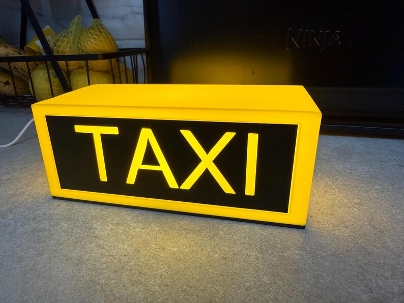 May include: A yellow rectangular taxi sign with the word "TAXI" in bold, illuminated letters. The sign has a black background and is lit from within, creating a bright, eye-catching display. The sign is on a gray surface.