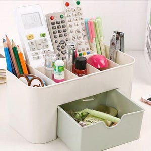 May include: A white desk organizer with multiple compartments and a pull-out drawer. The organizer holds various items, including pencils, pens, remote controls, and small bottles. A pink computer mouse and a stapler are also visible.