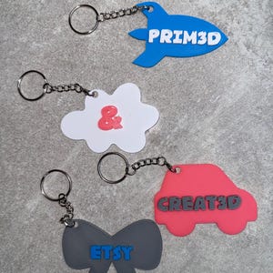 May include: Keychains in blue, white, gray, and pink, each with a silver chain and ring. The blue keychain is shaped like a rocket with the text "PRIM3D". A white cloud keychain has a red ampersand. A pink car keychain has "CREATE3D". A gray bow has "ETSY".