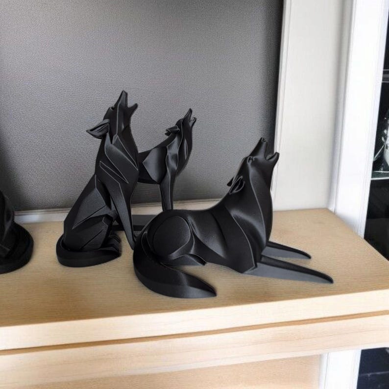May include: Three black wolf sculptures in various poses, howling upwards. The sculptures are on a light-colored wooden shelf against a gray wall. The wolves have a geometric, angular design.