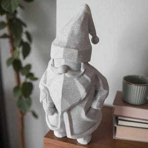 K&ouml;nnte beinhalten: Eine graue Zwergstatue mit spitzem Hut und langem Bart. Der Zwerg steht auf einem Holzregal. Eine gr&uuml;ne Pflanze und eine Tasse befinden sich im Hintergrund.