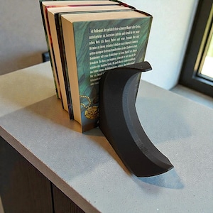 May include: A black, curved bookend supporting a stack of books. The bookend has a smooth, matte finish and a crescent shape. The books have green and gold covers with text in a foreign language.