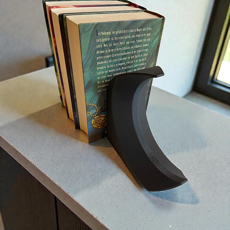 May include: A black, curved bookend supporting a stack of books. The bookend has a smooth, matte finish and a crescent shape. The books have green and gold covers with text in a foreign language.