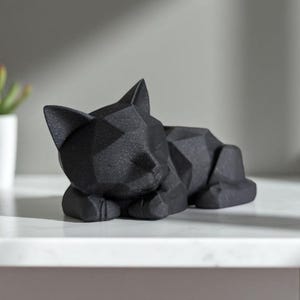 May include: A black geometric cat sculpture in a resting pose. The cat is lying down with its paws tucked beneath its body and its eyes closed. The sculpture has a faceted design, giving it a modern look. The cat is resting on a white surface.
