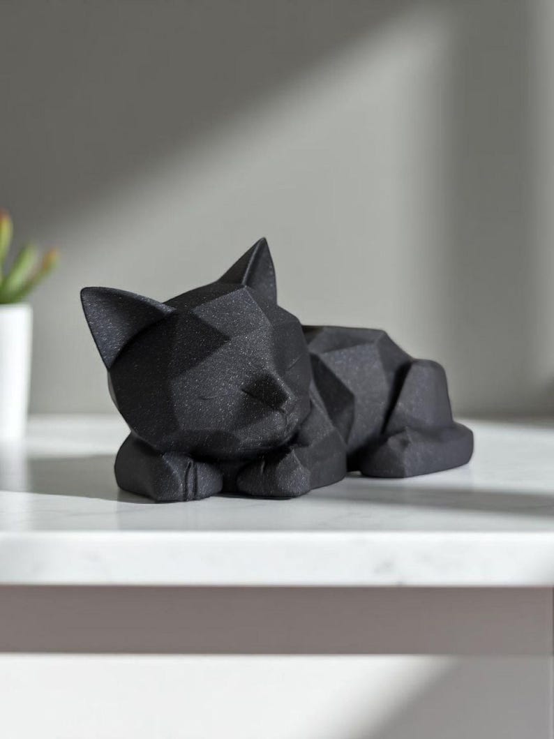 May include: A black geometric cat sculpture in a resting pose. The cat is lying down with its paws tucked beneath its body and its eyes closed. The sculpture has a faceted design, giving it a modern look. The cat is resting on a white surface.
