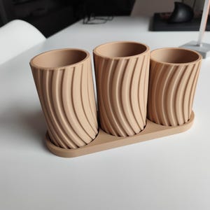 May include: A set of three beige cylindrical planters with a matching tray. Each planter has a textured, curved design. The planters are made of a solid material and are suitable for indoor or outdoor use.