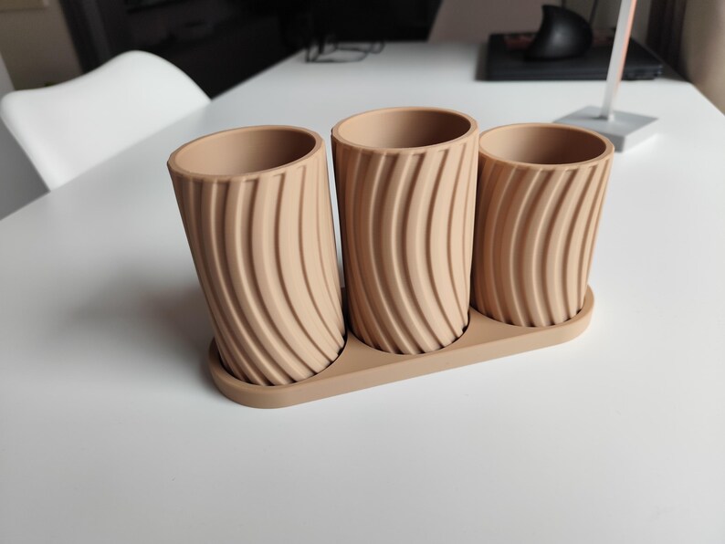 May include: A set of three beige cylindrical planters with a matching tray. Each planter has a textured, curved design. The planters are made of a solid material and are suitable for indoor or outdoor use.