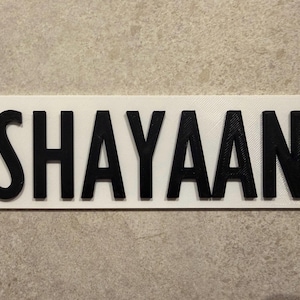 Pu&ograve; includere: Un cartello rettangolare bianco con il nome "SHAYAAN" in lettere nere in grassetto e in 3D. Il cartello &egrave; probabilmente una targhetta personalizzata o un oggetto decorativo, adatto per la cameretta di un bambino o come regalo personalizzato.