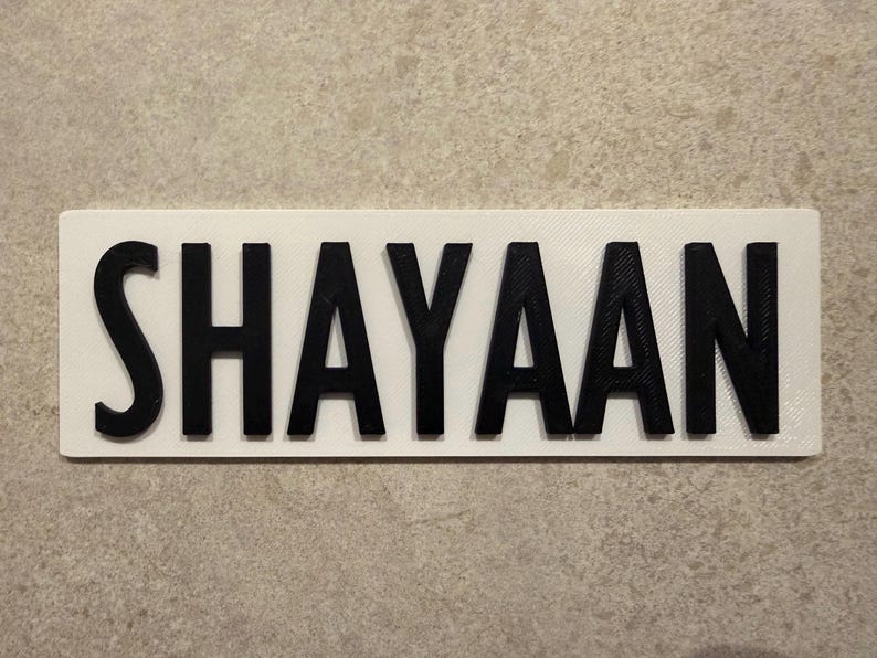 Pu&ograve; includere: Un cartello rettangolare bianco con il nome "SHAYAAN" in lettere nere in grassetto e in 3D. Il cartello &egrave; probabilmente una targhetta personalizzata o un oggetto decorativo, adatto per la cameretta di un bambino o come regalo personalizzato.