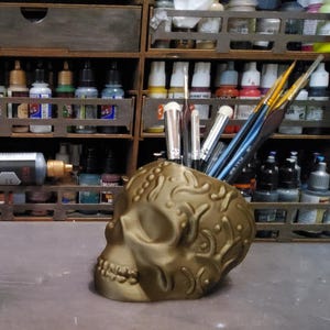 May include: A gold-colored skull-shaped container holding paintbrushes. The container has an ornate, embossed design. The background shows a wooden storage unit filled with various paint bottles, suggesting an artist's workspace.