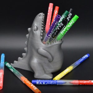 May include: A gray dinosaur-shaped pen holder with a wide open mouth, filled with colorful pens. Several pens are scattered around the base of the holder. The pens are various colors, including red, green, and blue.