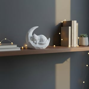 Peut inclure: Une figurine blanche repr&eacute;sentant un chat dormant sur un croissant de lune. La sculpture est pos&eacute;e sur une &eacute;tag&egrave;re en bois avec des livres et une petite plante en pot. Des guirlandes lumineuses d&eacute;coratives sont drap&eacute;es &agrave; proximit&eacute;.