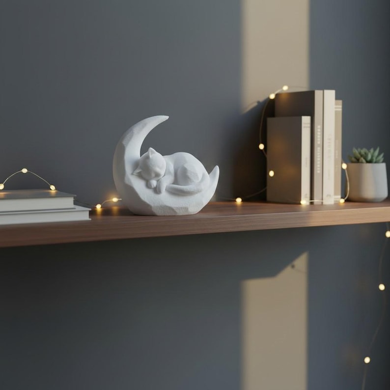 Peut inclure: Une figurine blanche repr&eacute;sentant un chat dormant sur un croissant de lune. La sculpture est pos&eacute;e sur une &eacute;tag&egrave;re en bois avec des livres et une petite plante en pot. Des guirlandes lumineuses d&eacute;coratives sont drap&eacute;es &agrave; proximit&eacute;.