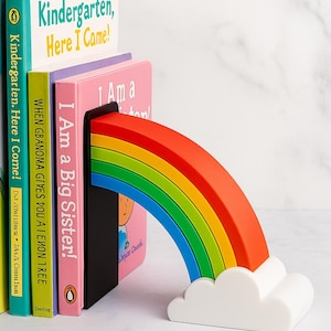 Puede incluir: Un sujetalibros de arco&iacute;ris colorido con una base de nube blanca. El arco&iacute;ris se extiende sobre una pila de libros infantiles, incluyendo "Kindergarten, Here I Came!" y "I Am a Big Sister!". Un elemento decorativo para la habitaci&oacute;n de un ni&ntilde;o.