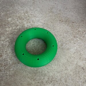 May include: A vibrant green ring with a series of small holes around its edge. The object, made from a solid material, is displayed on a neutral surface. The design is simple and geometric. The ring's diameter is approximately 10 cm.