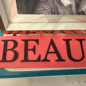 Pu&ograve; includere: Un cartello rettangolare color corallo con la parola "BEAU" in grassetto, lettere maiuscole nere. Il cartello &egrave; posto su una superficie azzurra, con un'immagine incorniciata sullo sfondo.