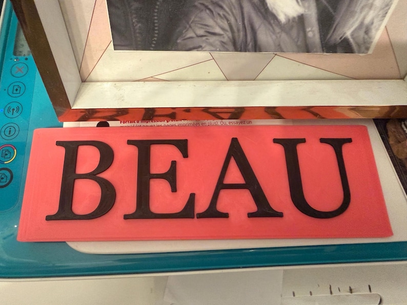Pu&ograve; includere: Un cartello rettangolare color corallo con la parola "BEAU" in grassetto, lettere maiuscole nere. Il cartello &egrave; posto su una superficie azzurra, con un'immagine incorniciata sullo sfondo.