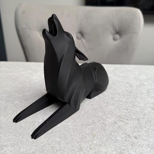 May include: A black, stylized sculpture of a howling wolf. The wolf is depicted in a reclining position with its head raised and mouth open. The sculpture has a modern, geometric design and is made of a matte black material.