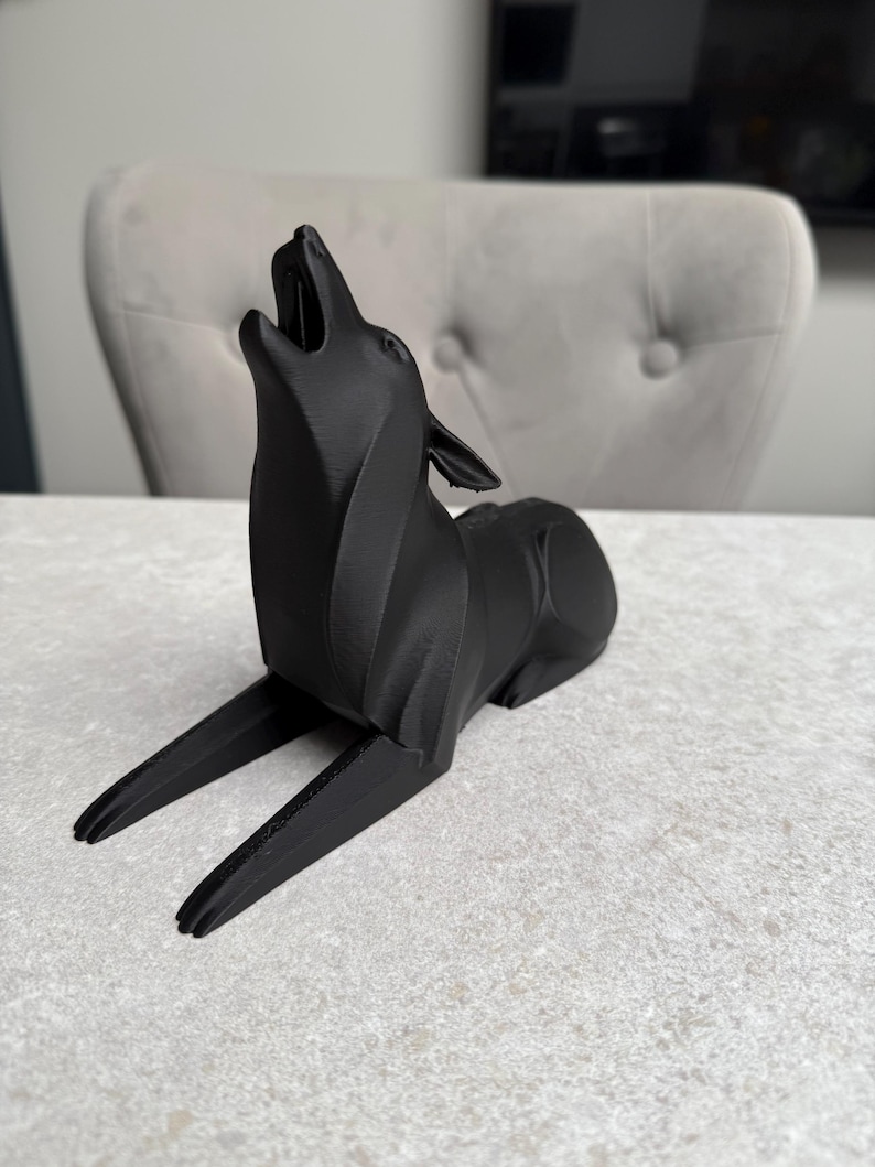 May include: A black, stylized sculpture of a howling wolf. The wolf is depicted in a reclining position with its head raised and mouth open. The sculpture has a modern, geometric design and is made of a matte black material.