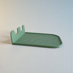Op de afbeelding: Een saliegroen, rechthoekig dienblad met een opstaande, gebogen rand en een houder met drie tanden. Het dienblad heeft een subtiel golfpatroon en is ontworpen om kleine items vast te houden. Het minimalistische ontwerp is geschikt voor diverse toepassingen.