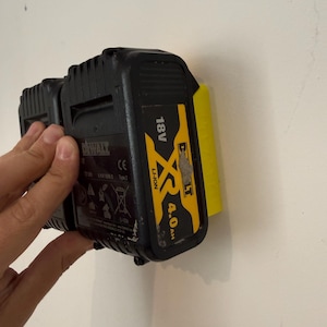 May include: Black and yellow DeWalt 18V XR 4.0 Ah Li-Ion battery pack. The battery is attached to a yellow holder on a white wall. The battery pack has the DeWalt logo and other markings.