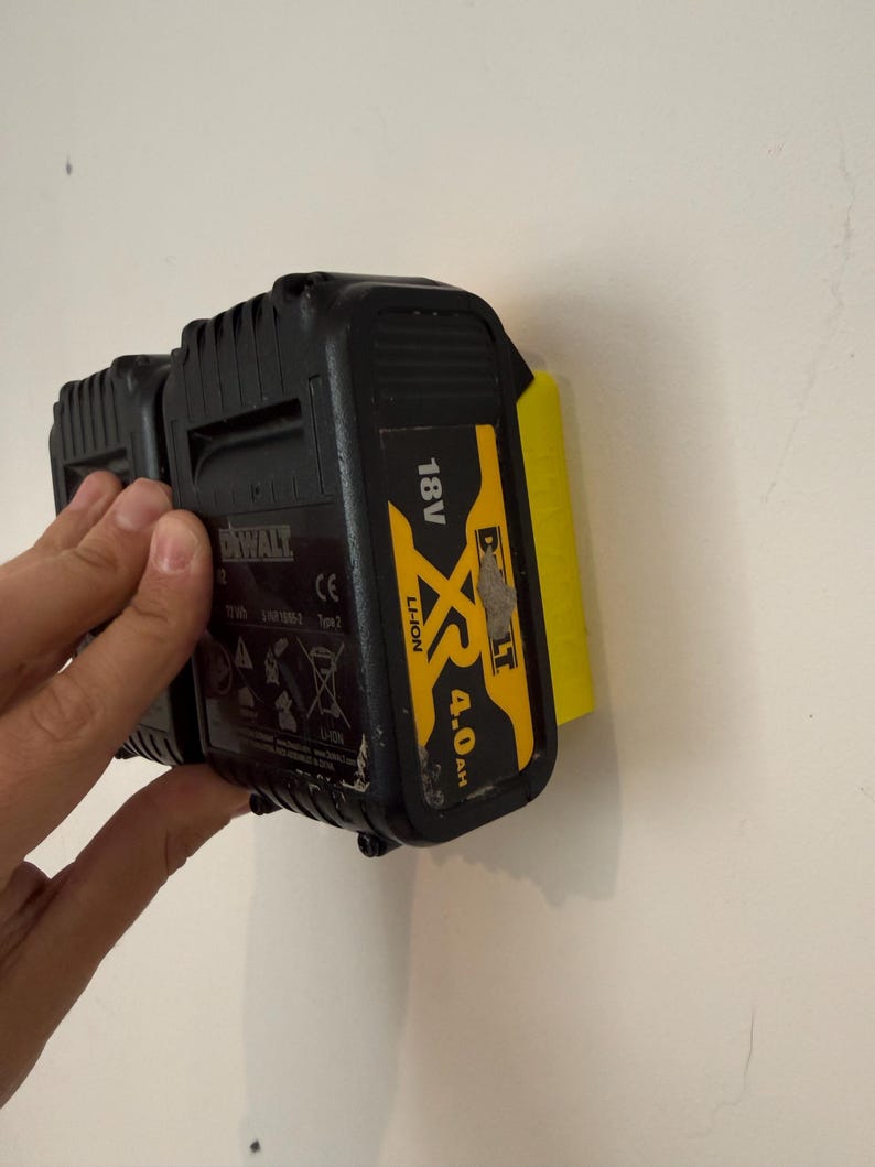 May include: Black and yellow DeWalt 18V XR 4.0 Ah Li-Ion battery pack. The battery is attached to a yellow holder on a white wall. The battery pack has the DeWalt logo and other markings.